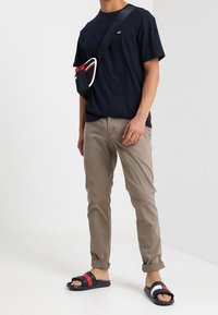 Granatowy t-shirt z okrągłym dekoltem, beżowe spodnie o dopasowanym kroju, czarne sandały z czerwonymi i białymi paskami oraz mała torba na ramię z czerwonymi akcentami.