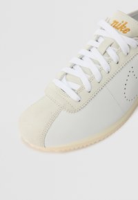 Gros plan sur une basket Nike beige et blanche avec des lacets blancs, un bout en daim, un swoosh perforé et un logo orange brodé sur la languette.