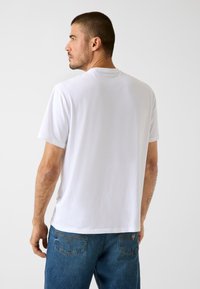 Weißes Baumwoll-T-Shirt mit rundem Ausschnitt und kurzen Ärmeln, kombiniert mit blauen Jeans aus Denim. Der Stoff wirkt glatt und leicht.