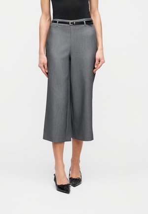 Donna che indossa pantaloni grigi a gamba larga, taglio cropped e vita alta con cintura nera e scarpe slingback nere con punta appuntita, in piedi davanti a uno sfondo bianco.