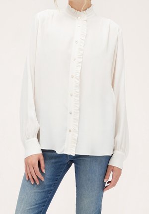 Witte blouse met lange mouwen, voorzien van een gerimpelde knoopsluiting en kraag, gedragen met een blauwe spijkerbroek, handen rustend langs de zijden.