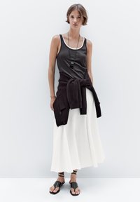 Débardeur gris avec bord blanc, longue jupe blanche, pull sombre noué à la taille, collier pendentif noir et sandales noires à lanières.