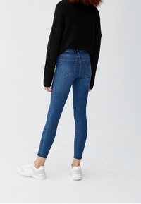 Persona vestida con jeans ajustados azules, suéter negro de manga larga y zapatillas blancas, de pie con la espalda hacia la cámara contra un fondo claro y liso.