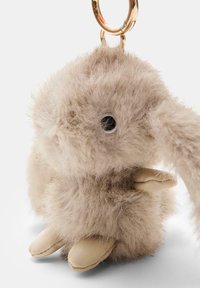 Porte-clés en forme de lapin beige duveteux avec une texture douce, de grandes oreilles, des yeux noirs et des pattes beiges en simili cuir, fixé à un anneau de clé en métal doré.