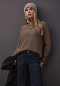 Brauner Strickpullover mit gerippten Bündchen, kombiniert mit dunkelblauen Jeans mit weitem Bein und oranger Naht. Das Model hält einen schwarzen Mantel.
