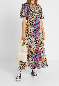 Frau in einem mehrfarbigen blumenartigen Patchworkkleid mit kurzen Ärmeln, die eine gewebte beige Tasche hält und schwarze High-Top-Sneaker trägt.