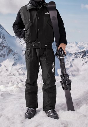 Osoba ubrana w czarny strój narciarski i buty, trzymająca czarną nartę na ośnieżonej górze z ośnieżonymi szczytami w tle.