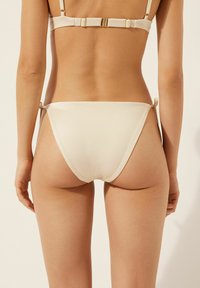 Modell sedd bakifrån iförd en ljusbeige bikini med remmar och knytningar, mot en neutral bakgrund. Huden är solbrun och len.