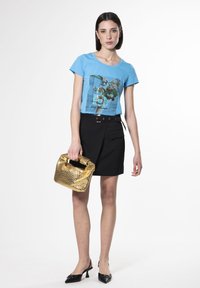 Camiseta azul de manga corta con diseño gráfico, combinada con una falda negra y sosteniendo un bolso dorado brillante. Zapatos de tacón negro tipo slingback.