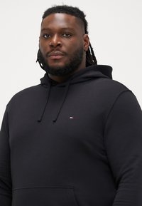 Czarna bawełniana bluza z kapturem, z kieszenią typu kangur, regulowanym kapturem na sznurki oraz małym logo z niebieskimi, białymi i czerwonymi akcentami na klatce piersiowej.