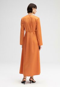 Oranje jurk met lange mouwen in een gladde stof, met een getailleerde taille en een zwierige rok. Zwarte hak sandalen maken de outfit compleet.