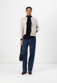 Polstret beige jakke med horisontalt quiltet mønster, glidelås og høy krage, kombinert med en svart turtleneck og vide denimjeans.