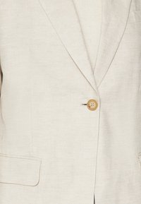 Béžový lněný sako s jedním hnědým knoflíkem, klopovým límcem a dvěma předními kapsami. Měkká textura a upravený design.