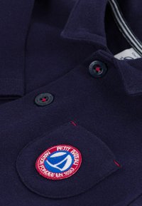 Mørk marineblå polo skjorte med et fremtredende brodert logo på brystlommen, to sorte knapper og kontrastfarge rød stitching detalj.