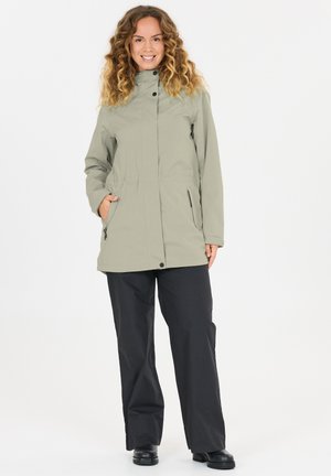 Kvinde med krøllet hår iført lys grøn jakke med lommer, sorte bukser og sorte sko, stående og smilende mod hvid baggrund.