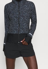 Černá zipová bunda s modrým tečkovaným vzorem, vysokým límcem a otvory na palce; v kombinaci s černými šortkami. Lehký, sportovní design.