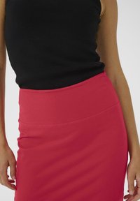 Jupe crayon rouge extensible avec une taille haute et une texture lisse, associée à un débardeur noir ajusté. Accent sur la zone des hanches et de la taille.