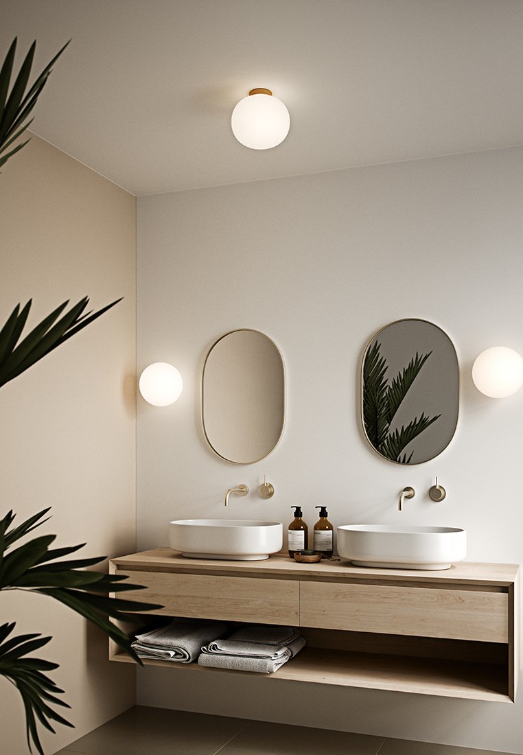 Weiße Keramiksäulen auf einem Holztisch, mit zwei ovalen Spiegeln. Messingarmaturen, Wandleuchte und dunkle Flaschen auf der Ablage. Strukturierte Handtücher darunter.