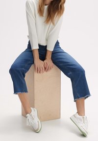 Personne portant des baskets blanches, un jean bleu effiloché et un haut blanc côtelé à manches longues, assise sur une boîte en bois, les mains posées devant.