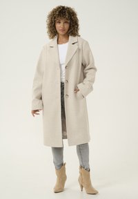 Manteau en mélanger de laine beige clair avec un col cranté, deux boutons, poches latérales et coupe décontractée, associé à un jean gris et des bottines beige.