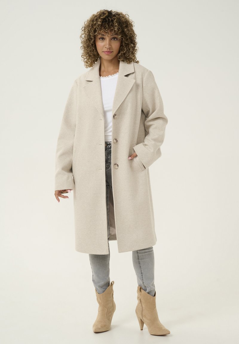 Manteau en mélanger de laine beige clair avec un col cranté, deux boutons, poches latérales et coupe décontractée, associé à un jean gris et des bottines beige.
