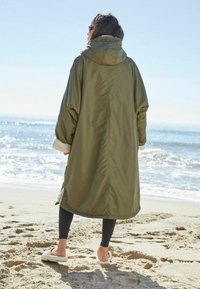 Olivengrøn vandtæt frakke med hætte, der har en blød cremelining, oversize ærmer og en afslappet pasform, båret over sorte leggings.