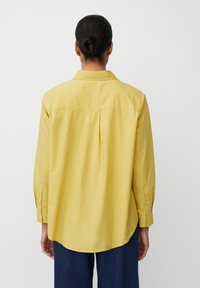 Camicia gialla a maniche lunghe con orlo curvato, piega posteriore e polsini con bottoni, realizzata in morbido cotone. Nessun motivo o accento visibile.