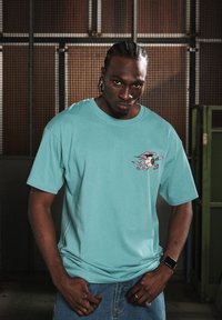 Camiseta de algodón turquesa con mangas cortas que presenta un diseño de dibujo animado en rosa en el lado izquierdo del pecho. Modelo vistiendo un par de jeans de mezclilla.