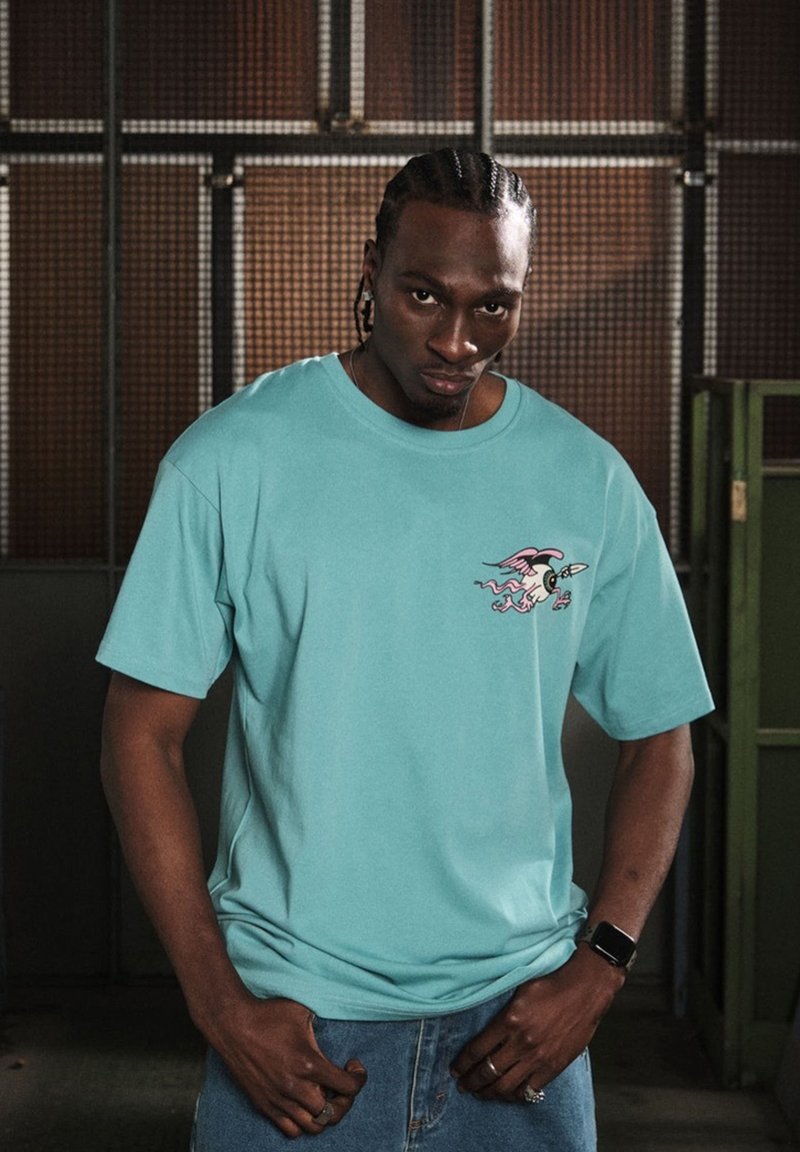 Camiseta de algodón turquesa con mangas cortas que presenta un diseño de dibujo animado en rosa en el lado izquierdo del pecho. Modelo vistiendo un par de jeans de mezclilla.
