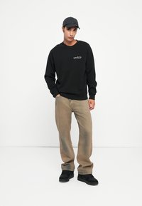 Melnā kapuce ar izšūtu logotipu krūšu daļā, apvienota ar gaiši brūnām biksēm un melnu apaviem. Modelis, kas valkā atbilstošu cepuri.