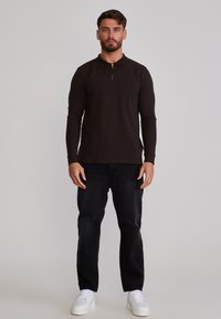 Bărbat în picioare, cu fața înainte, purtând un polo negru cu mâneci lungi, pantaloni negri și adidași albi, pe un fundal simplu.