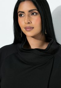 Czarny strój z drapowanym dekoltem i gładką teksturą. Modelka ma długie czarne włosy i nosi złote kolczyki wiszące. Delikatne oświetlenie podkreśla rysy twarzy.