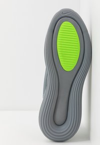 Szara podeszwa sportowego buta z charakterystycznym wzorem i zielonym akcentem, z wyraźnymi rowkami zapewniającymi przyczepność i elastyczność.