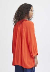 Blusa arancione comoda con maniche a tre quarti, dettaglio plissettato sul retro e polsini abbottonati, realizzata in un tessuto leggero e testurizzato.