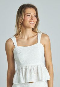Top peplum bianco con scollatura quadrata e spalline larghe. Presenta un design floreale ricamato su un tessuto testurizzato.