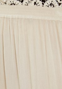 Falda plisada de color beige claro con un tejido texturizado. Cuenta con una cinturilla ajustada y un delicado borde de encaje en la parte superior.