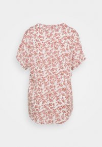 Blouse blanche à manches courtes, coupe ample, avec un motif floral rouge pâle couvrant l'ensemble, présentée sur un fond uni.