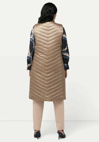 Dlouhá zlatá péřová vesta s chevronovým prošíváním, v kombinaci s vzorovanou modro-krémovou košilí, béžovými kalhotami a námořnickými lodičkami.