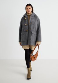 Manteau duffle en laine gris avec des boutonnières en bois et des poches, associé à une robe pull beige et des collants noirs. Sac à bandoulière marron et bottes vertes.