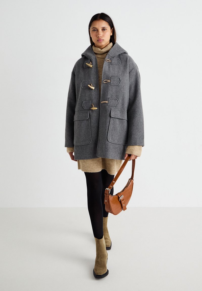 Szary wełniany płaszcz typu duffle z drewnianymi zapięciami i kieszeniami, w połączeniu z beżową sukienką swetrową i czarnymi rajstopami. Brązowa torba na ramię i zielone buty.