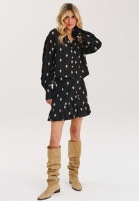 Černá blůza se zlatým květinovým vzorem, doplněná k volné sukni s volány. Modelka má na sobě uvolněné béžové boty a stojí s jednou rukou na boku.