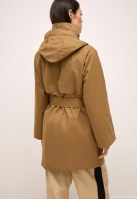 Femme vue de dos portant un manteau beige à capuche avec ceinture et un pantalon crème avec une bande latérale noire sur un fond clair.