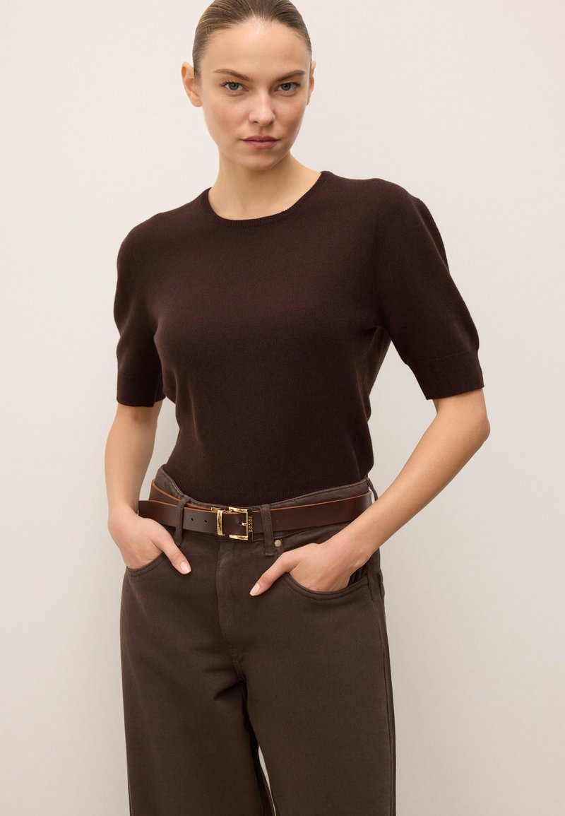 Barna rövid ujjú pulóver, magas derekú barna nadrággal párosítva, amelyhez barna öv tartozik, arany színű csattal.