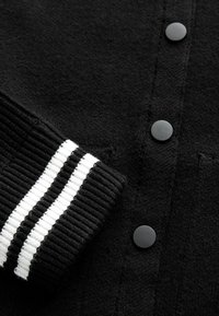 Black cardigan featuring a textured knit, five black buttons, and a ribbed cuff with white stripes.
