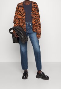 Pomarańczowy i czarny kardigan w paski tygrysie o lekkiej teksturze, noszony na ciemnym t-shircie i niebieskich dżinsach. Czarne skórzane buty i kurtka.