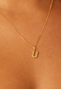Gouden ketting met een U-vormige hanger met gegraveerde bloemmotieven, hangend aan een dunne, getextureerde ketting tegen een huidkleurige achtergrond.