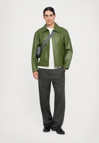 Grüne Lederjacke, weißes T-Shirt, graue, plissierte Hose, schwarze Schuhe und eine kleine schwarze Umhängetasche. Hat einen Kragenstil.