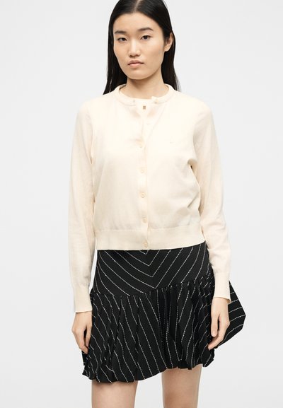 Νεαρή γυναίκα που φοράει μια μπεζ ζακέτα με κουμπιά και μακριά μανίκια, και μια μαύρη φούστα με λευκές διαγώνιες ρίγες και βολάν.