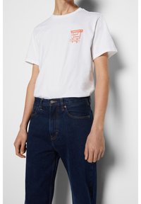 Vit bomullströja med korta ärmar, med ett orange grafiskt tryck på vänster bröst. I kombination med mörkblå denimjeans.