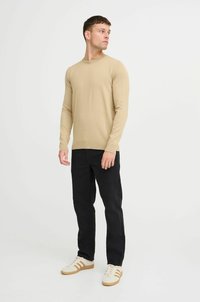 Suéter beige claro hecho de un suave tejido de punto, con cuello redondo y mangas largas, combinado con pantalones negros y zapatillas blancas.